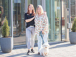 Eine jüngere und eine ältere Frau, Tochter und Mutter, stehen mit einem kleinen Hund an der Leine vor dem Eingang der Pflegeeinrichtung in Albersloh.